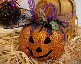 May include: Three orange glitter pumpkins with black and purple accents. Each pumpkin has a different facial expression. The pumpkins are sitting on a bed of straw.