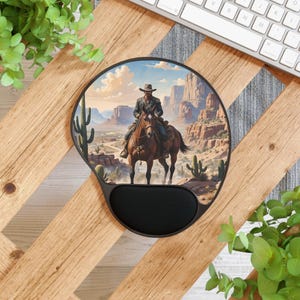 May include: Round black mouse pad with a wrist rest. The pad features a detailed illustration of a cowboy on horseback in a desert landscape with cacti and mesas. The image is framed by a black border.
