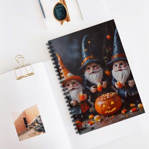 Może przedstawiać: Spiralny notatnik z ilustracją o tematyce Halloween. Obraz przedstawia trzy gnomy w czapach czarownic, trzymające cukierki w kształcie kukurydzy i zebrane wokół wyrzeźbionej dyni. Gnomy mają długie białe brody i ubrane są w czarne kurtki.