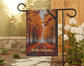 May include: A garden flag featuring a fall scene with a tree-lined road and the text "Hello Autumn" in gold. The trees display vibrant orange and red foliage, creating a seasonal aesthetic. The flag is displayed on a black metal stand.