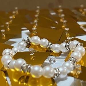 May include: A white pearl bracelet with silver accents. The bracelet is made of round pearls and silver beads. The bracelet is lying on a gold surface.