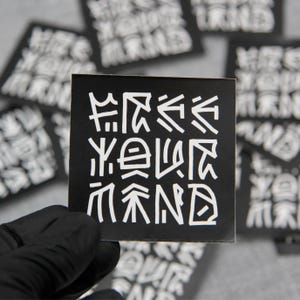 May include: Square black sticker with white stylized text that reads "Free Your Mind". The sticker is held by a gloved hand, with other similar stickers in the background. The design is modern and graphic.