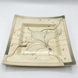 May include: A rectangular ceramic serving tray with a raised rim and a stylised floral design in shades of blue, brown, and white. The tray has a textured surface and a slightly curved shape.
