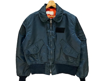 May include: A dark teal bomber jacket with a black collar, cuffs, and waistband. The jacket has two front pockets, a black patch on the chest, and a zipper closure. The jacket is hanging on a wooden hanger.
