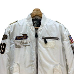 Pu&ograve; includere: Giacca bomber bianca con chiusura a zip, con toppe e la scritta "SIMPSON". La giacca ha una toppa con la bandiera americana e il numero "69" sulla manica. Il testo sulla giacca recita "LA NOSTRA LUNGA EREDIT&Agrave; DI INNOVAZIONE NELLA SICUREZZA".