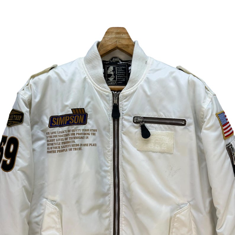 Pu&ograve; includere: Giacca bomber bianca con chiusura a zip, con toppe e la scritta "SIMPSON". La giacca ha una toppa con la bandiera americana e il numero "69" sulla manica. Il testo sulla giacca recita "LA NOSTRA LUNGA EREDIT&Agrave; DI INNOVAZIONE NELLA SICUREZZA".