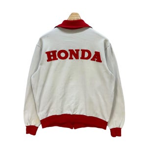 Può includere: Una giacca bianca con zip, bordi rossi e un grande logo "HONDA" rosso sul retro.