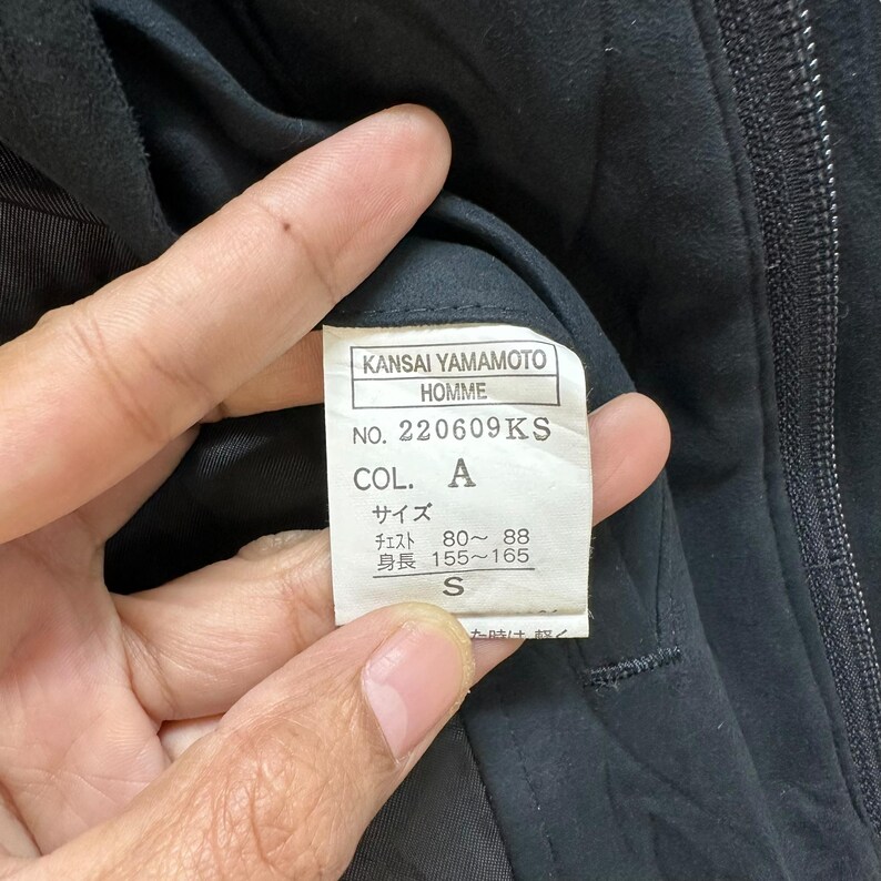 Może przedstawiać: Czarna metka odzieżowa z białym tekstem, na kt&oacute;rej widnieje napis "KANSAI YAMAMOTO HOMME NO. 220609KS COL. A サイズ チェスト 80~88 身長 155~165 S"