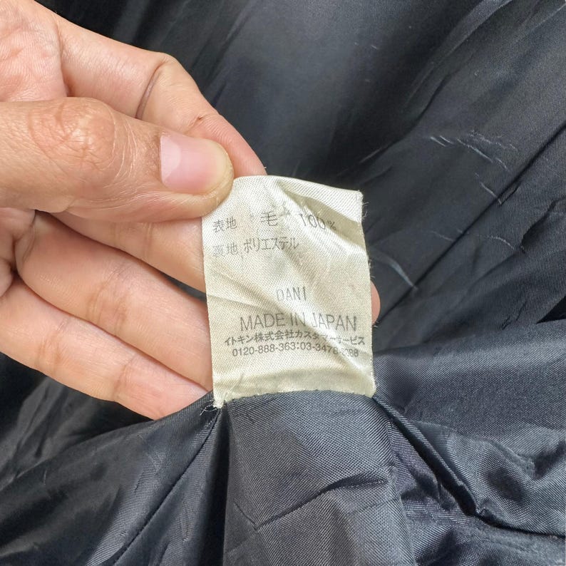 Peut inclure: Gros plan sur une &eacute;tiquette de v&ecirc;tement tenue par une main. L'&eacute;tiquette est blanc cass&eacute; avec du texte japonais et les mots "MADE IN JAPAN". L'&eacute;tiquette indique &eacute;galement les mati&egrave;res : laine et polyester.