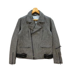 Peut inclure: Veste en laine grise avec fermeture éclair asymétrique noire et détails en cuir noir. La veste présente un col cranté, plusieurs poches zippées et des sangles réglables avec boucles à la taille. Accrochée sur un cintre en bois.