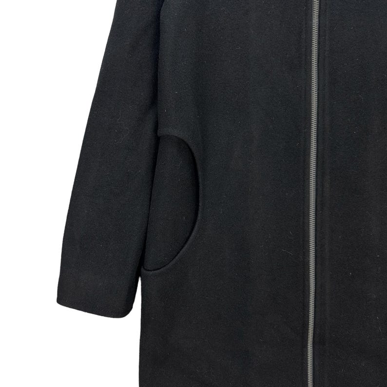 Peut inclure: Gros plan d'un manteau noir avec une fermeture &eacute;clair et une grande poche. La manche est visible, et le tissu semble &ecirc;tre une mati&egrave;re douce, semblable &agrave; de la laine. Le manteau est de couleur noire unie.