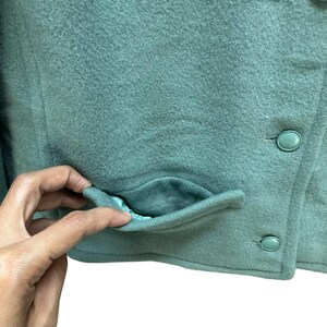 May include: A teal green jacket with a single pocket and two buttons. The jacket is made of a soft, fuzzy material.