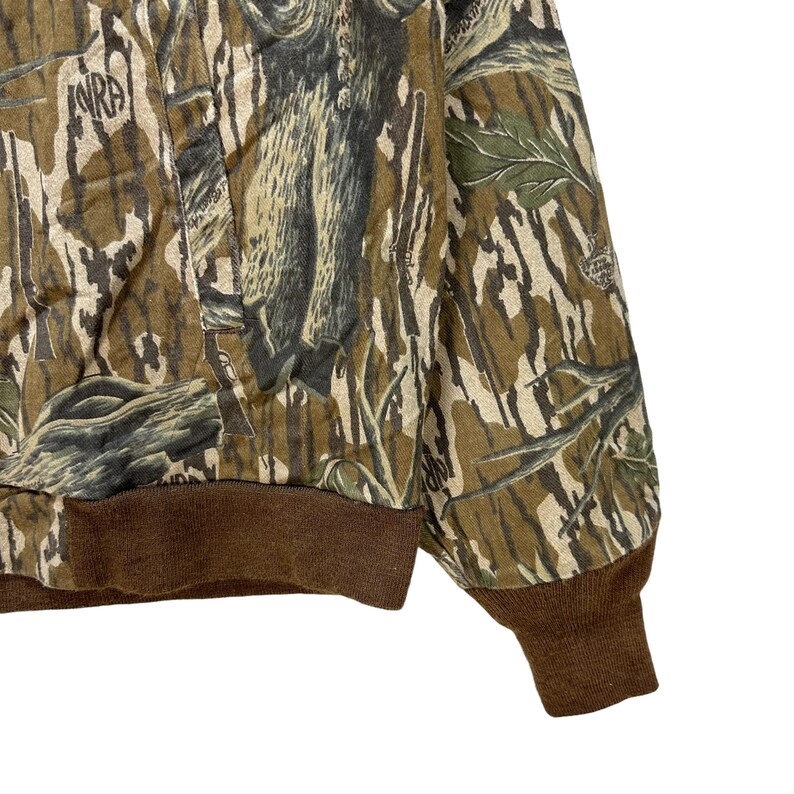 May include: A camouflage-patterned jacket with brown ribbed cuffs and waistband. The jacket features a mix of green, brown, and tan colors, creating a woodland camouflage design. The letters "NRA" are visible on the jacket.