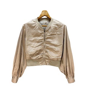 May include: A beige bomber jacket with a ruched front and a ribbed hem and cuffs. The jacket has a zip-up closure and long sleeves. The jacket is hanging on a wooden hanger against a white background.