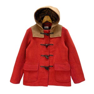 Könnte beinhalten: Ein roter Woll-Dufflecoat mit brauner Kapuze und Knebelverschlüssen. Der Mantel hat zwei große Taschen vorne und ein Etikett mit der Aufschrift "CAUCHO."