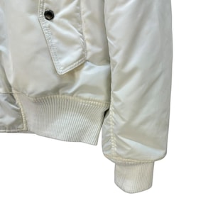 Pu&ograve; includere: Giacca bomber color crema con polsini e orlo a coste. La giacca presenta una tasca frontale e una chiusura con bottoni. La manica ha un polsino a coste. La giacca &egrave; realizzata in un materiale liscio, possibilmente nylon o poliestere.