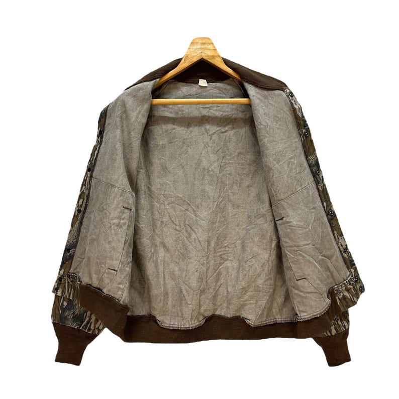 May include: A brown and green camouflage jacket with a brown collar and cuffs, hanging on a wooden hanger. The jacket is open, revealing a light brown interior lining. The jacket has a classic bomber style.
