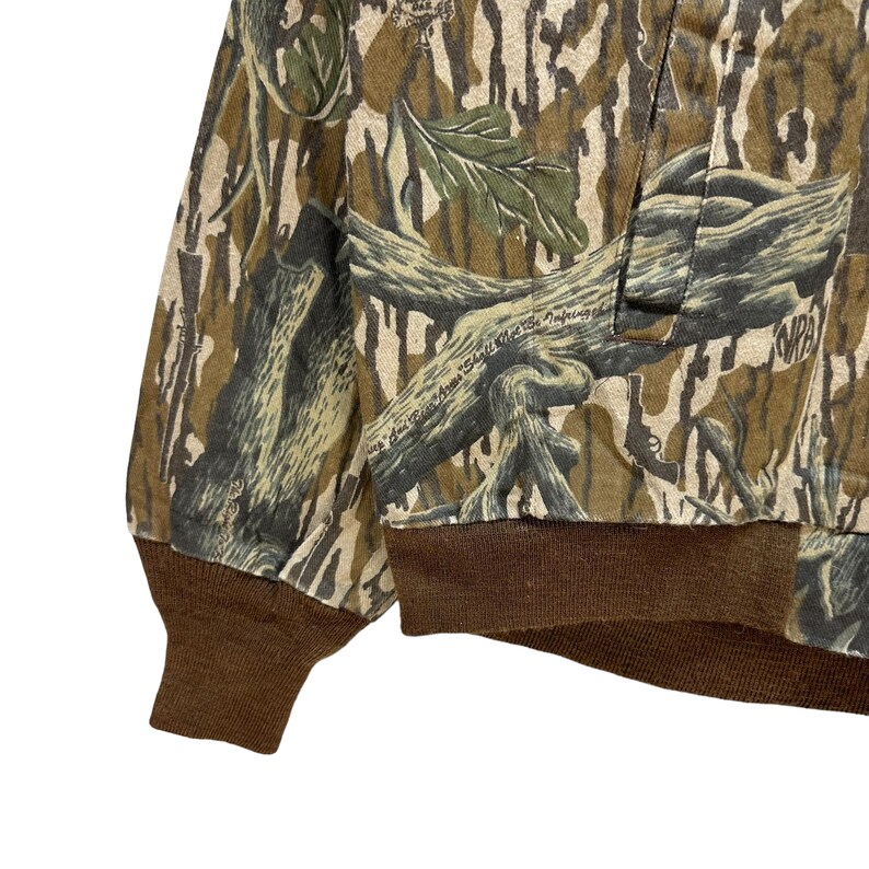 May include: Camouflage-patterned jacket with brown ribbed cuffs and hem. The jacket features a woodland camouflage design in shades of green, brown, and beige. The text "NRA" is visible on the jacket. The jacket is made of a durable fabric.