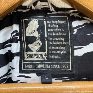 Pu&ograve; includere: Primo piano dell'etichetta di una giacca da moto in bianco e nero. L'etichetta presenta il logo "SIMPSON" e il testo "NORTH CAROLINA SINCE 1959". La giacca ha un motivo in bianco e nero.