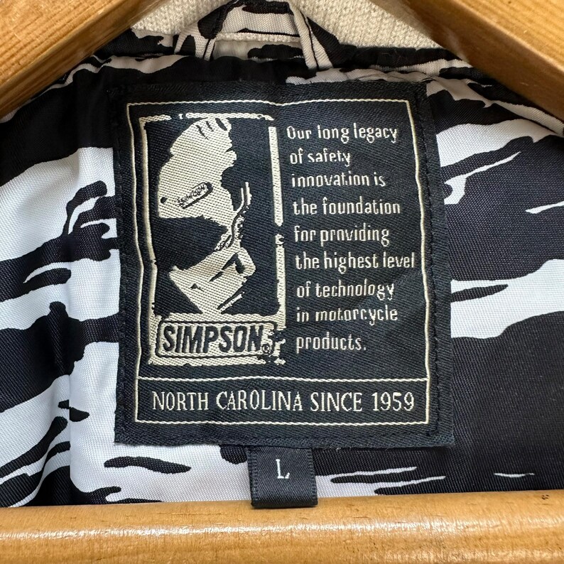 Pu&ograve; includere: Primo piano dell'etichetta di una giacca da moto in bianco e nero. L'etichetta presenta il logo "SIMPSON" e il testo "NORTH CAROLINA SINCE 1959". La giacca ha un motivo in bianco e nero.