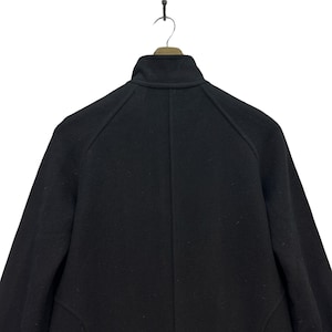 Peut inclure: Un manteau en laine noir, vu de dos, suspendu &agrave; un cintre en bois. Le manteau pr&eacute;sente un col montant et une coupe ajust&eacute;e. L'image est prise sur fond blanc, mettant en valeur les d&eacute;tails et la texture du manteau.