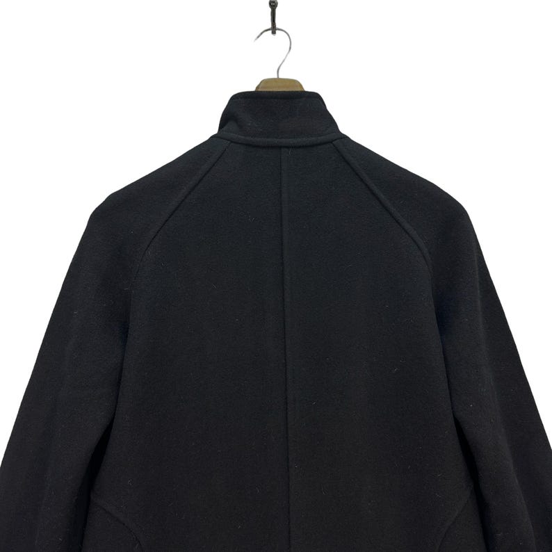 Peut inclure: Un manteau en laine noir, vu de dos, suspendu &agrave; un cintre en bois. Le manteau pr&eacute;sente un col montant et une coupe ajust&eacute;e. L'image est prise sur fond blanc, mettant en valeur les d&eacute;tails et la texture du manteau.