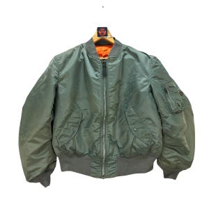 May include: Olive green bomber jacket with a gray ribbed collar, cuffs, and waistband. The jacket features a zipper closure, two front pockets, and a small pocket on the left sleeve. The interior lining is bright orange.