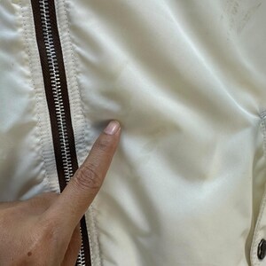 Pu&ograve; includere: Primo piano di una giacca color crema con cerniera e finiture marroni. Il tessuto della giacca appare liscio e leggermente lucido. Un dito tocca la superficie della giacca.