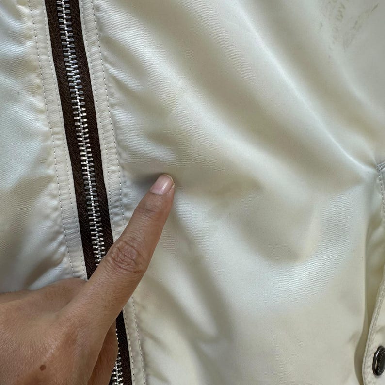 Pu&ograve; includere: Primo piano di una giacca color crema con cerniera e finiture marroni. Il tessuto della giacca appare liscio e leggermente lucido. Un dito tocca la superficie della giacca.