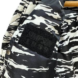 Pu&ograve; includere: Primo piano di una giacca a fantasia bianca e nera. La giacca presenta un design simile al camouflage con forme irregolari. Una toppa in velcro nera &egrave; visibile all'interno della giacca. La giacca &egrave; appesa a una gruccia di legno.