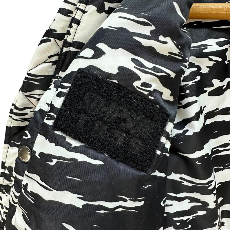 Pu&ograve; includere: Primo piano di una giacca a fantasia bianca e nera. La giacca presenta un design simile al camouflage con forme irregolari. Una toppa in velcro nera &egrave; visibile all'interno della giacca. La giacca &egrave; appesa a una gruccia di legno.