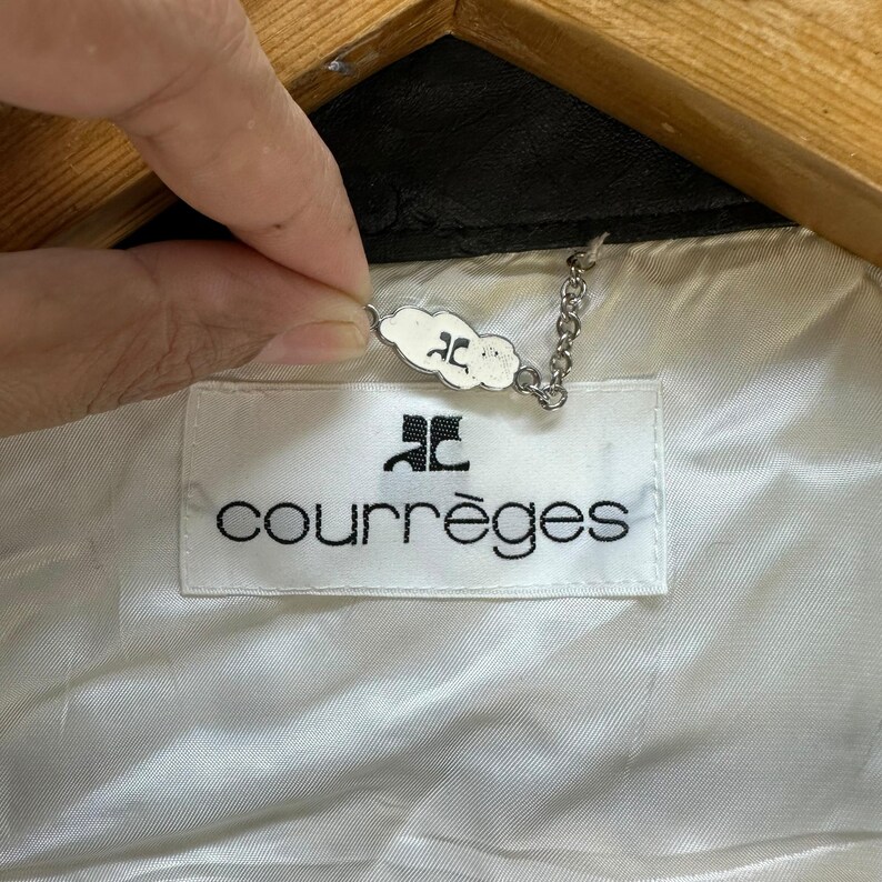 May include: A white label with the text "Courr&egrave;ges" in black lettering and a small silver tag with the letter "C" in a cloud shape. The label is attached to a black leather jacket.