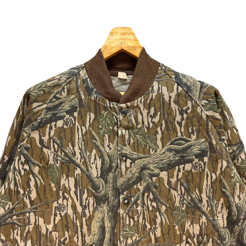 May include: Camouflage bomber jacket with a brown collar and button closure. The jacket features a tree branch pattern in shades of brown, green, and tan. The letters "NRA" are visible on the jacket.