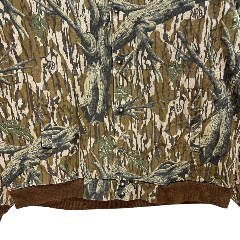 May include: Camouflage jacket with a brown ribbed hem and cuffs. The jacket features a button-up front and a pattern of tree branches and leaves in shades of brown, green, and tan. The letters "NRA" are visible on the jacket.