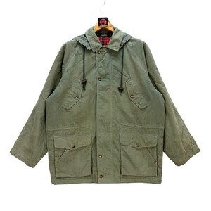 Può includere: Giacca con cappuccio verde oliva con due ampie tasche frontali e chiusure con bottoni. La giacca ha un cappuccio con coulisse e una fodera a quadri visibile sul colletto. È appesa a una gruccia di legno.