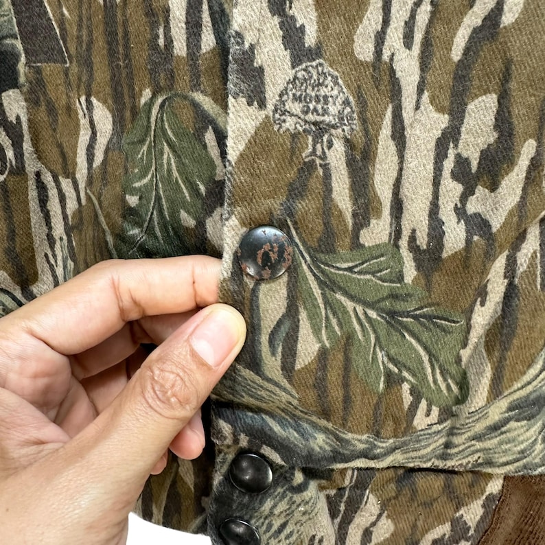 May include: Close-up of a camouflage jacket with a button closure. The jacket features a green, brown, and tan leaf pattern. The "Mossy Oak" logo is visible on the jacket. The jacket is made of a textured fabric.