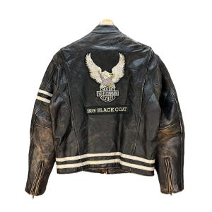 Peut inclure: Une veste en cuir noir avec un grand écusson brodé d'un aigle au dos portant l'inscription MOTOR EAGLES COMMANDO CYCLES et BIG BLACK COAT. La veste présente des bandes horizontales blanches sur les manches et la taille, ainsi que des poignets zippés.