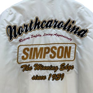 Pu&ograve; includere: Giacca color crema con scritte ricamate. La parola "Northcarolina" &egrave; arcuata nella parte superiore in corsivo nero e oro. Sotto c'&egrave; "SIMPSON" in un rettangolo dorato. Il testo "The Winning Edge since 1959" &egrave; sotto.
