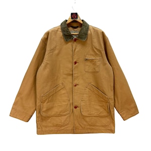 Op de afbeelding: Een bruin werkjack met een groene corduroy kraag. De jas heeft vier voorzakken, een knoopsluiting en een kleine borstzak. Het hangt aan een hanger.
