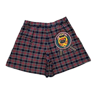 May include: Red and blue plaid shorts with a black cat patch on the right side. The patch has a crown and the text "New Kid" on it.