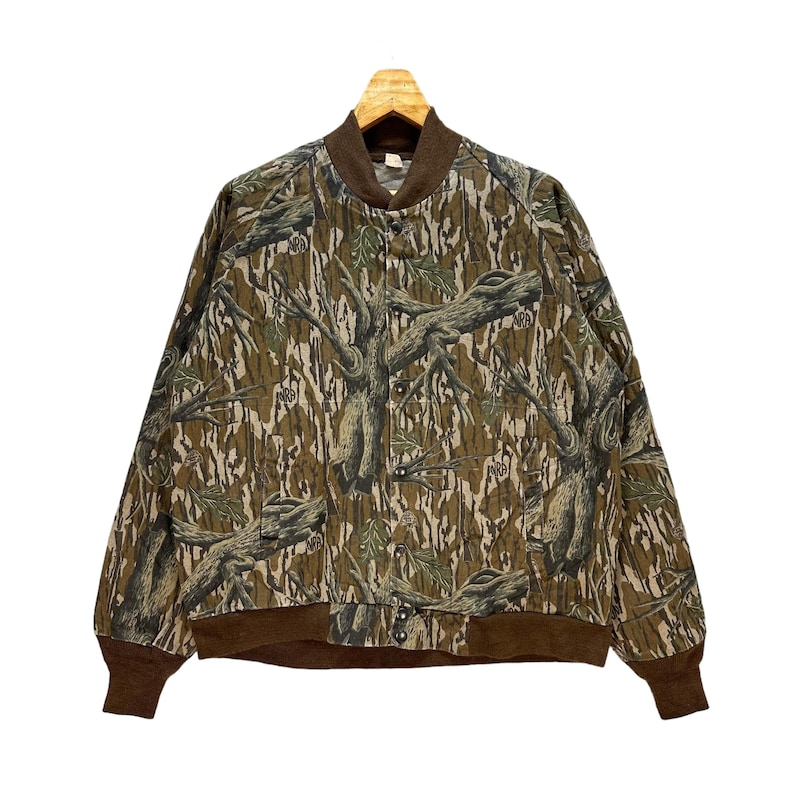 May include: A button-up bomber jacket with a camouflage pattern of brown, green, and tan tree branches and leaves. The jacket has a brown collar, cuffs, and waistband. The jacket is hanging on a wooden hanger.