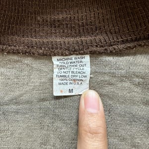 May include: Close-up of a brown garment's label with care instructions and the text "MACHINE WASH COLD WATER. TURN INSIDE OUT GENTLE CYCLE DO NOT BLEACH TUMBLE DRY LOW 100% COTTON MADE IN U.S.A M".