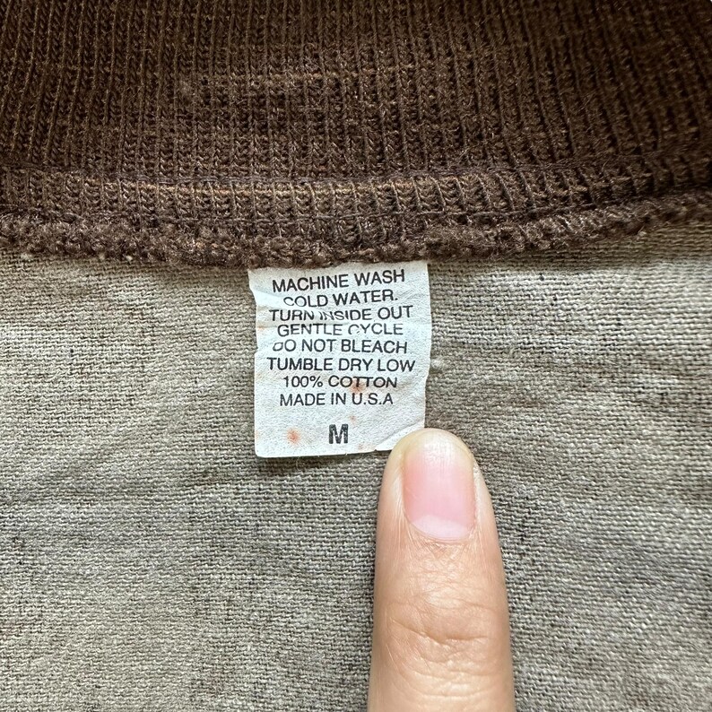 May include: Close-up of a brown garment's label with care instructions and the text "MACHINE WASH COLD WATER. TURN INSIDE OUT GENTLE CYCLE DO NOT BLEACH TUMBLE DRY LOW 100% COTTON MADE IN U.S.A M".