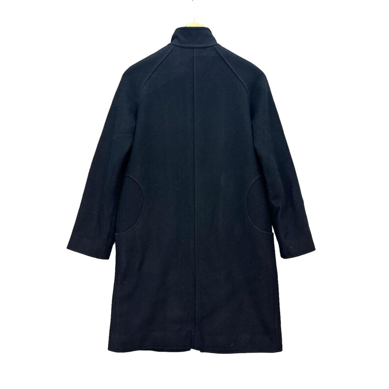 Peut inclure: Un manteau en laine noir avec un col montant et des manches longues. Le manteau a deux grandes poches arrondies et une coupe ajust&eacute;e. Il est suspendu &agrave; un cintre en bois sur fond blanc.
