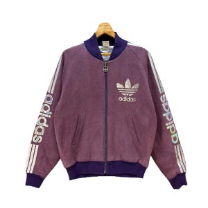 Può includere: Giacca Adidas vintage viola con zip, logo argentato sul petto e nome Adidas stampato sulle maniche. La giacca ha un corpo viola con finiture blu navy su colletto, polsini e orlo. Sono presenti due tasche anteriori.