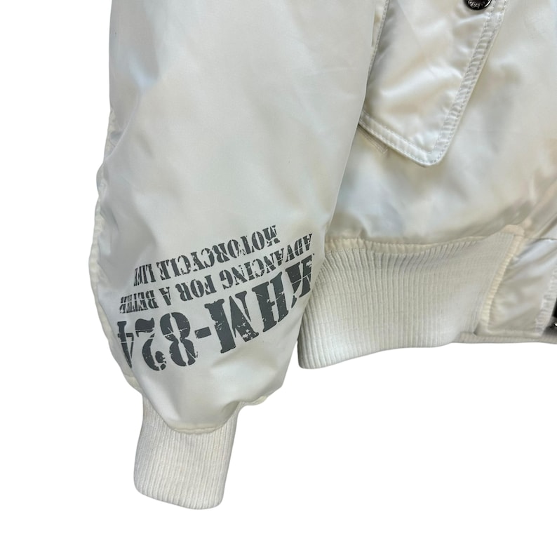 Pu&ograve; includere: Giacca da moto bianco sporco con polsini e orlo a coste. La manica sinistra presenta la scritta "KHM-824 ADVANCING FOR A BETTER MOTORCYCLE LIFE" in nero. La giacca ha una texture trapuntata e un classico stile bomber.