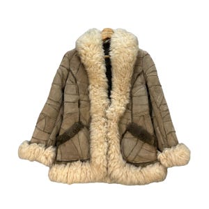 May include: A tan patchwork coat with a cream-colored shearling collar, cuffs, and trim. The coat has two pockets and is hanging on a wooden hanger. The coat is made of suede and shearling.