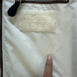 Pu&ograve; includere: Primo piano dell'interno di una giacca. La fodera &egrave; bianco sporco, con una cerniera marrone e un tirazip in pelle nera. &Egrave; visibile una toppa rettangolare con testo in rilievo. La giacca sembra vintage.