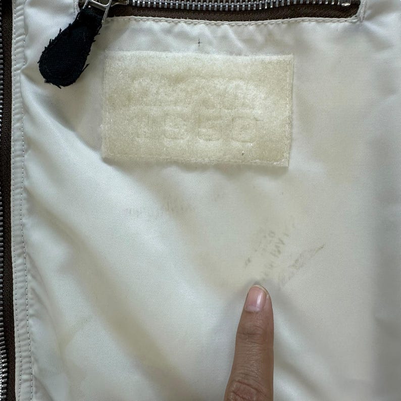 Pu&ograve; includere: Primo piano dell'interno di una giacca. La fodera &egrave; bianco sporco, con una cerniera marrone e un tirazip in pelle nera. &Egrave; visibile una toppa rettangolare con testo in rilievo. La giacca sembra vintage.