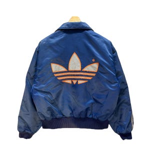 Può includere: Una giacca bomber vintage Adidas blu con un grande logo Adidas bianco e arancione sul retro.